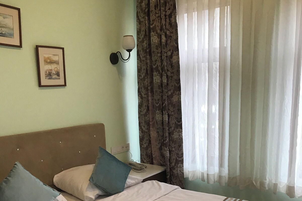A neatly made bed with pillows, a striped blanket, and towels on the bed. There is a nightstand with a lamp and a phone. The room has light green walls, a window with curtains, and framed pictures on the wall.