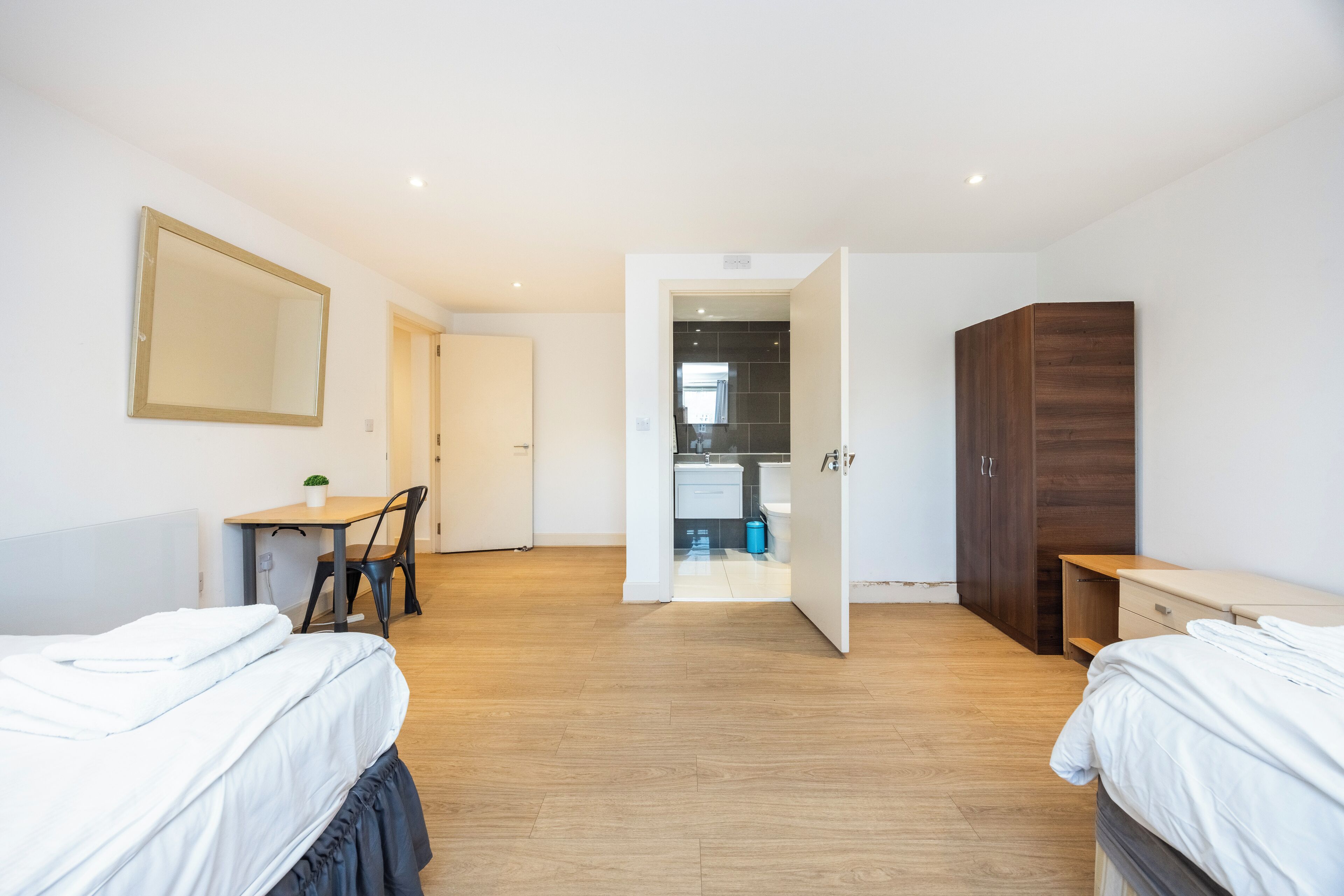 A room with two beds, a desk, a chair, a wardrobe, and a bathroom visible through an open door.