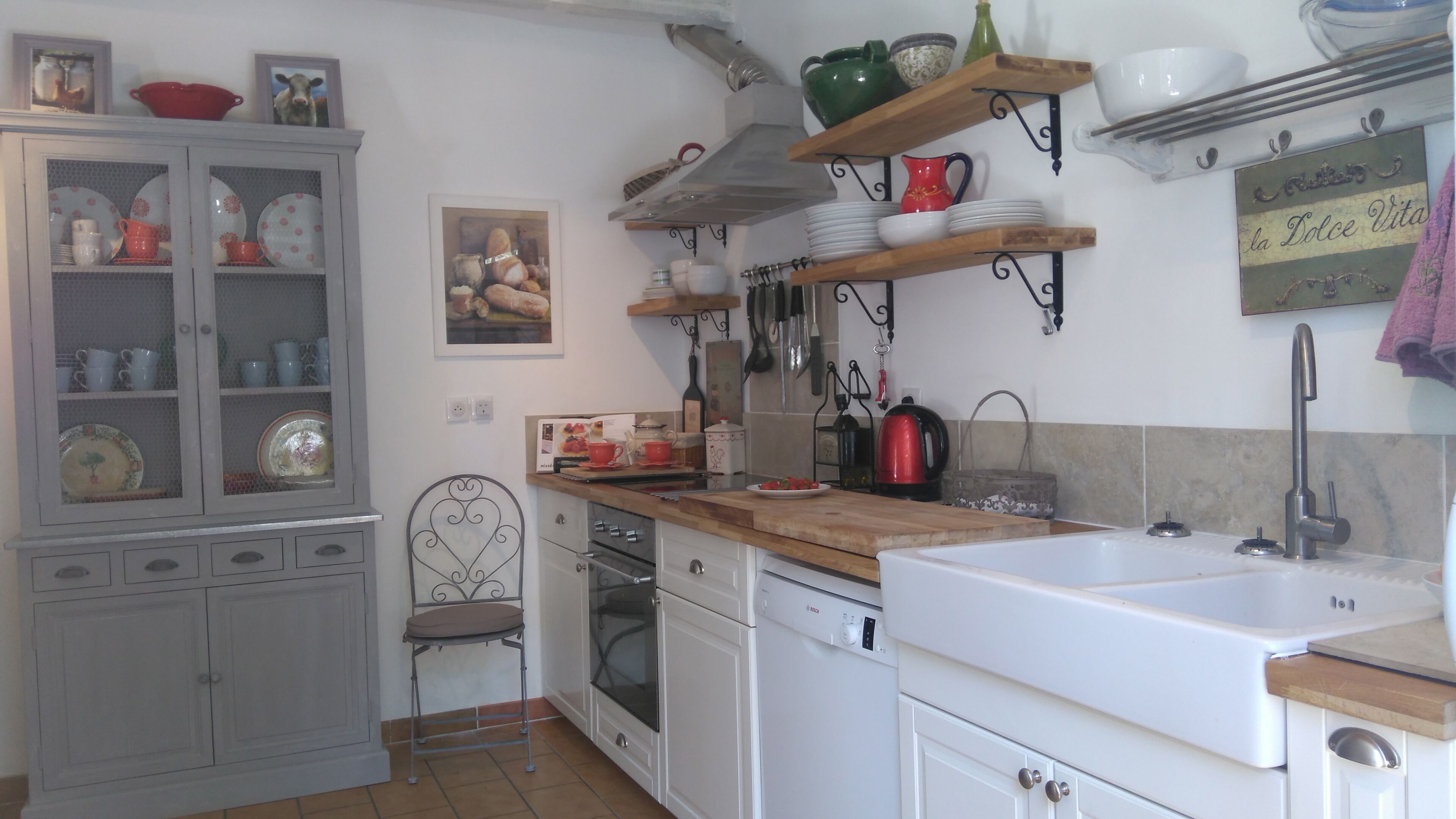 A kitchen with white cabinets, a wooden countertop, a sink, and a stove. There are shelves above the countertop holding dishes and a kettle.