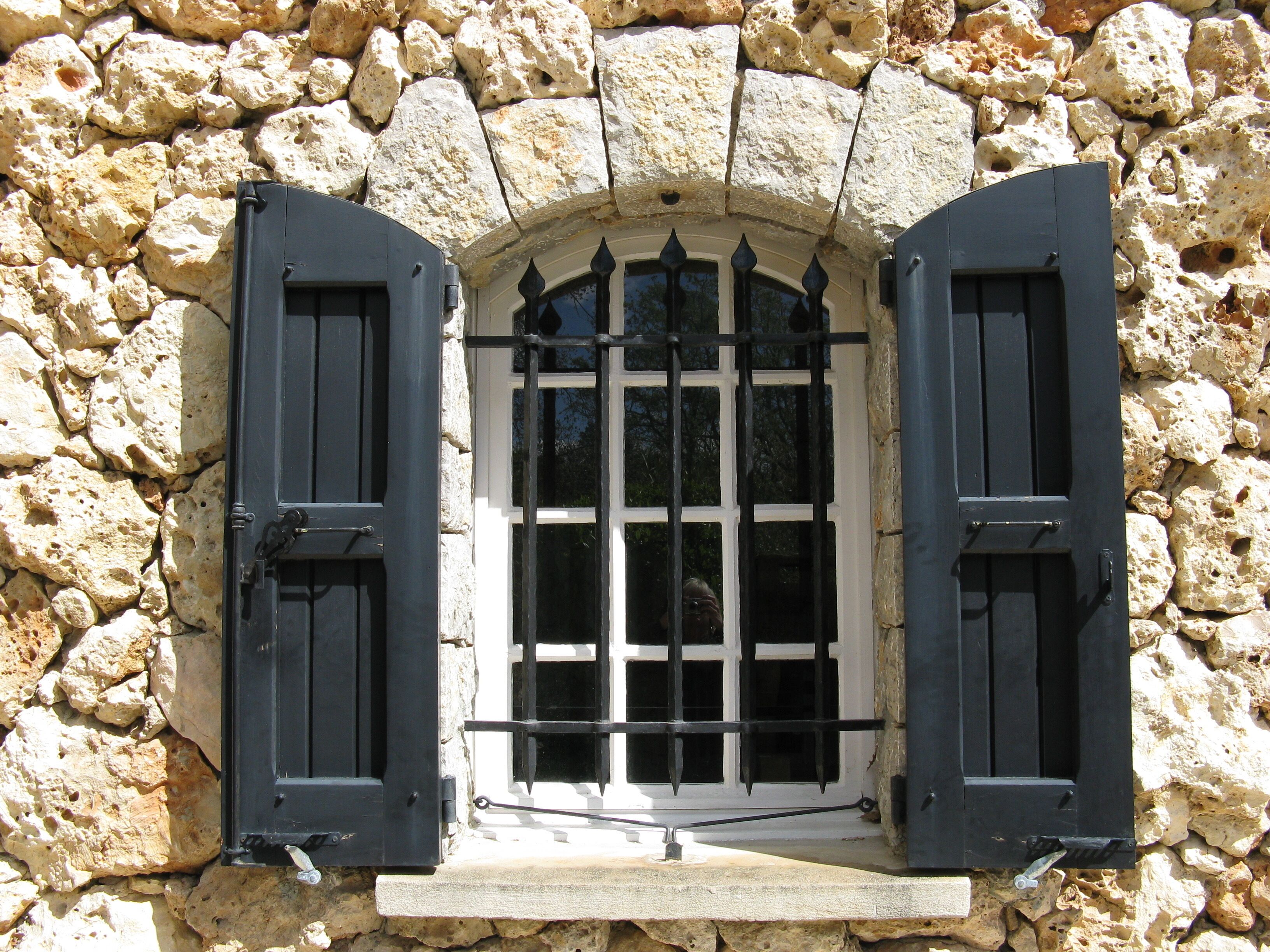 A stone wall with a window featuring black shutters and a metal grille.