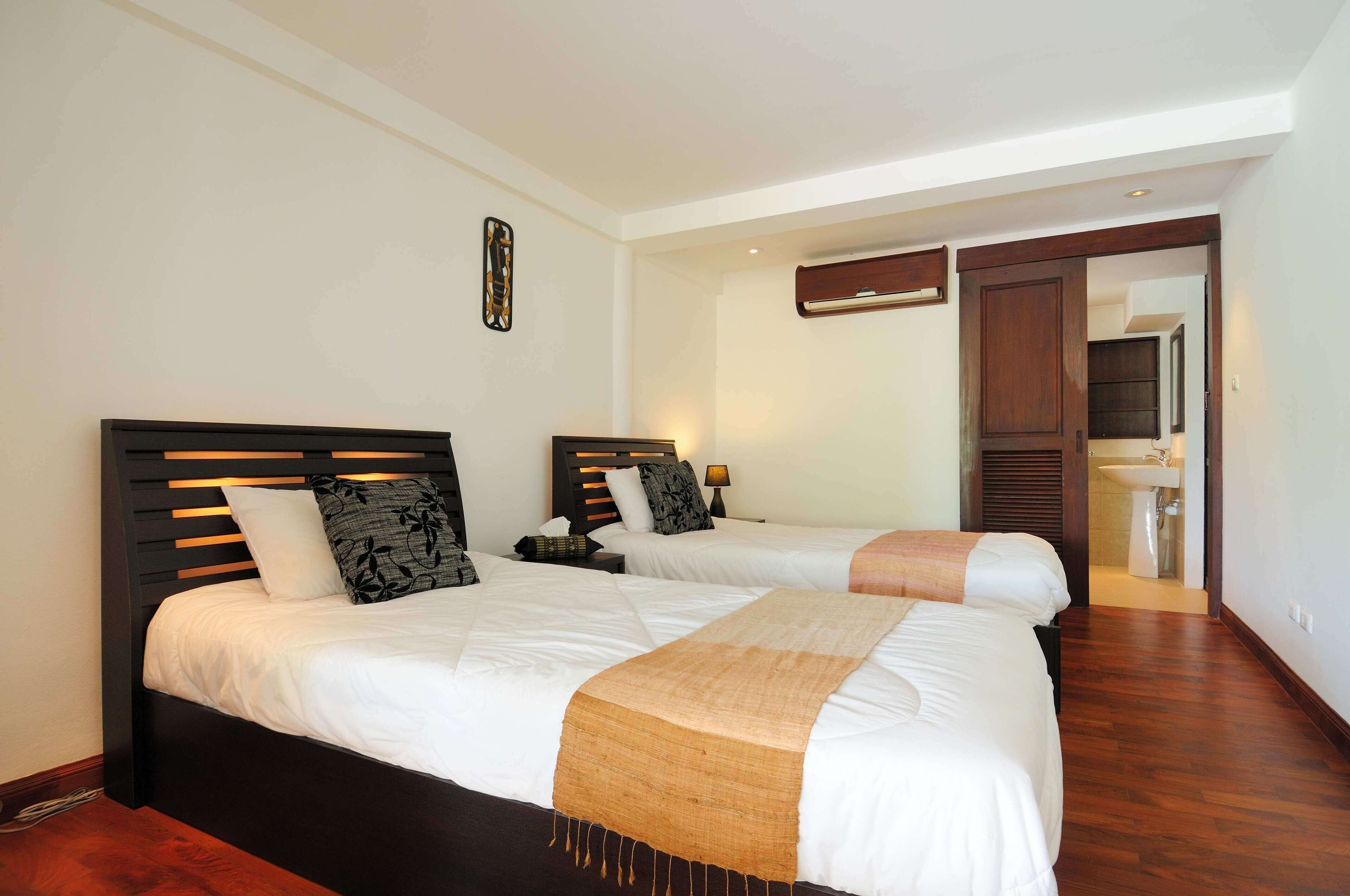 A hotel room with two beds, a wooden headboard, a bedside table with a lamp, and a bathroom visible through an open door.