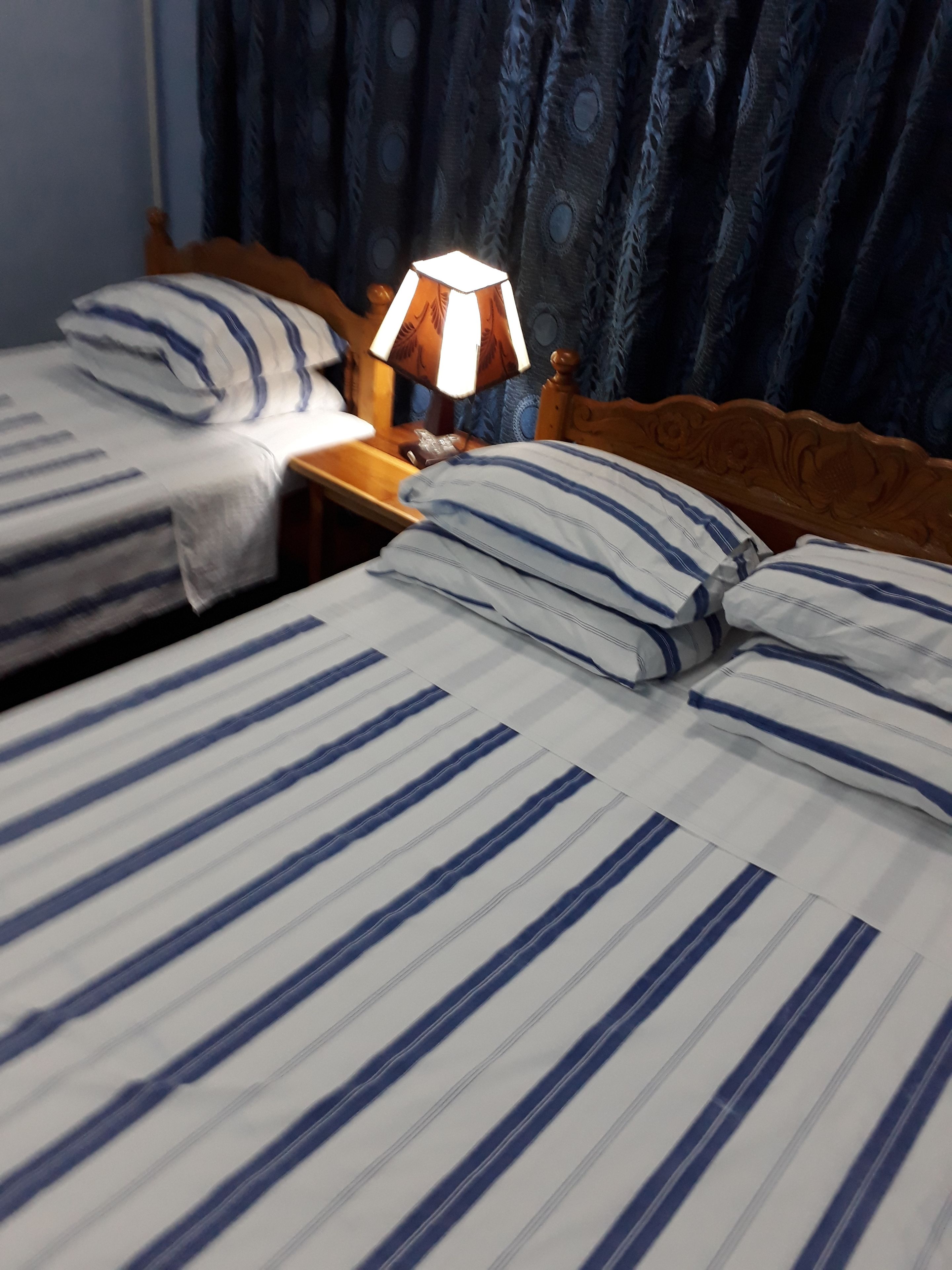 A hotel room with two beds, each with striped bedding and matching pillows. There is a wooden headboard, a nightstand with a lamp, and a curtain with a circular pattern.