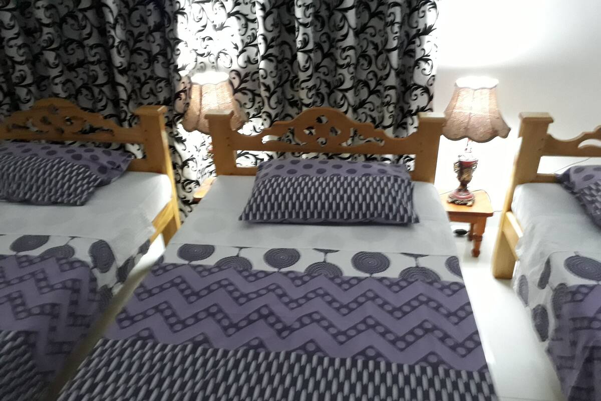 A room with two wooden beds, each with a patterned bedspread and pillows. There are two bedside tables with lamps. The room has patterned curtains.