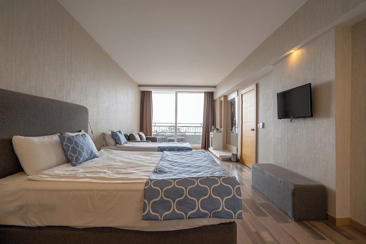 A hotel room with a large bed, a bench, a TV, and a balcony with curtains.