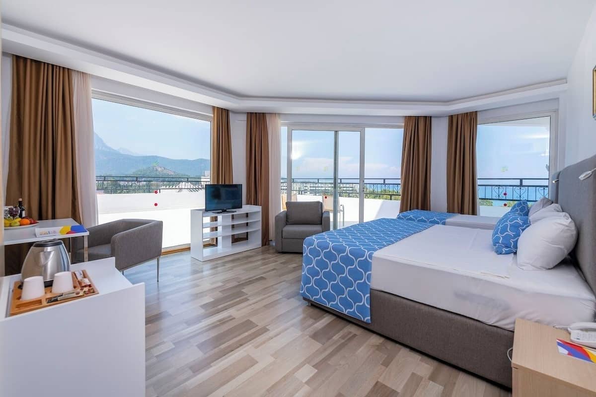 A hotel room with a large bed, a desk, a chair, and a TV. The room has large windows with a view of the sea and mountains.