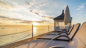 Ett soligt däck med solstolar och en liten paviljong med utsikt över havet vid solnedgången.