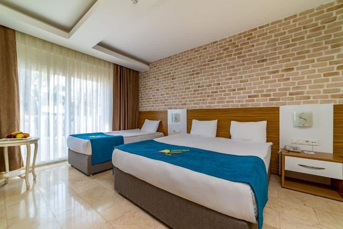 A hotel room with two beds, a brick wall, a window with curtains, a bedside table, and a small table with fruit.