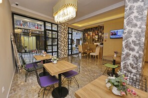 Un hall de hotel con zona de comedor, mesas de madera y sillas moradas, un candelabro y una pantalla de televisión.