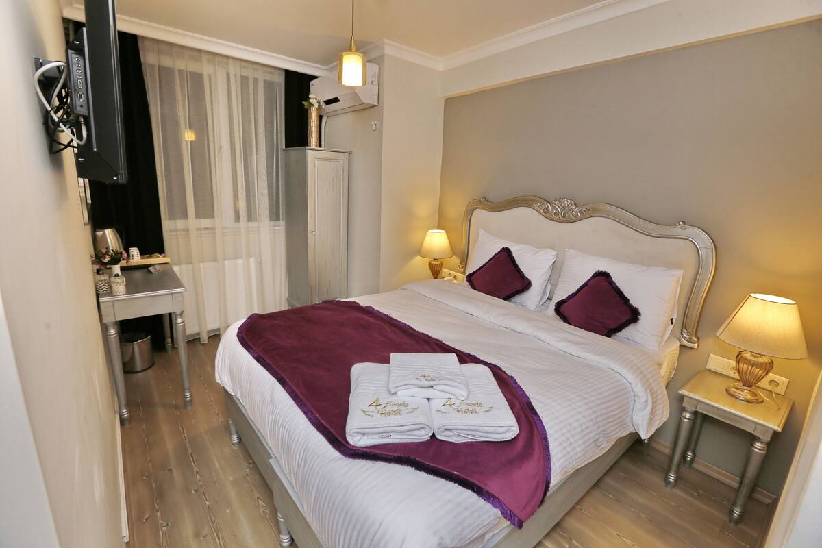 Una cama bien hecha con un edredón morado, dos toallas blancas y un cabecero con detalles decorativos.