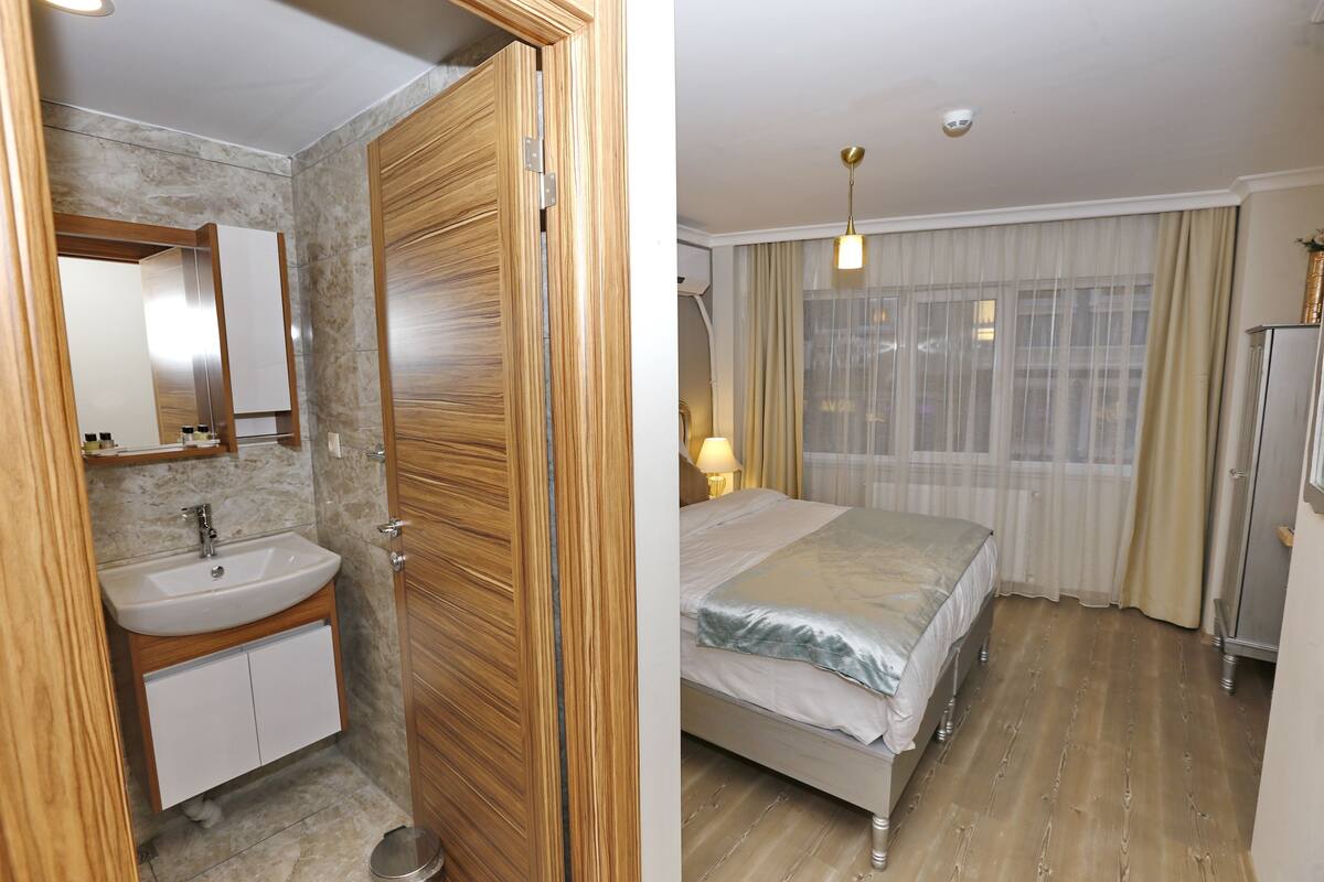 Un baño con tocador de madera, lavamanos y espejo. La habitación tiene suelo de madera y un ventanal con cortinas.