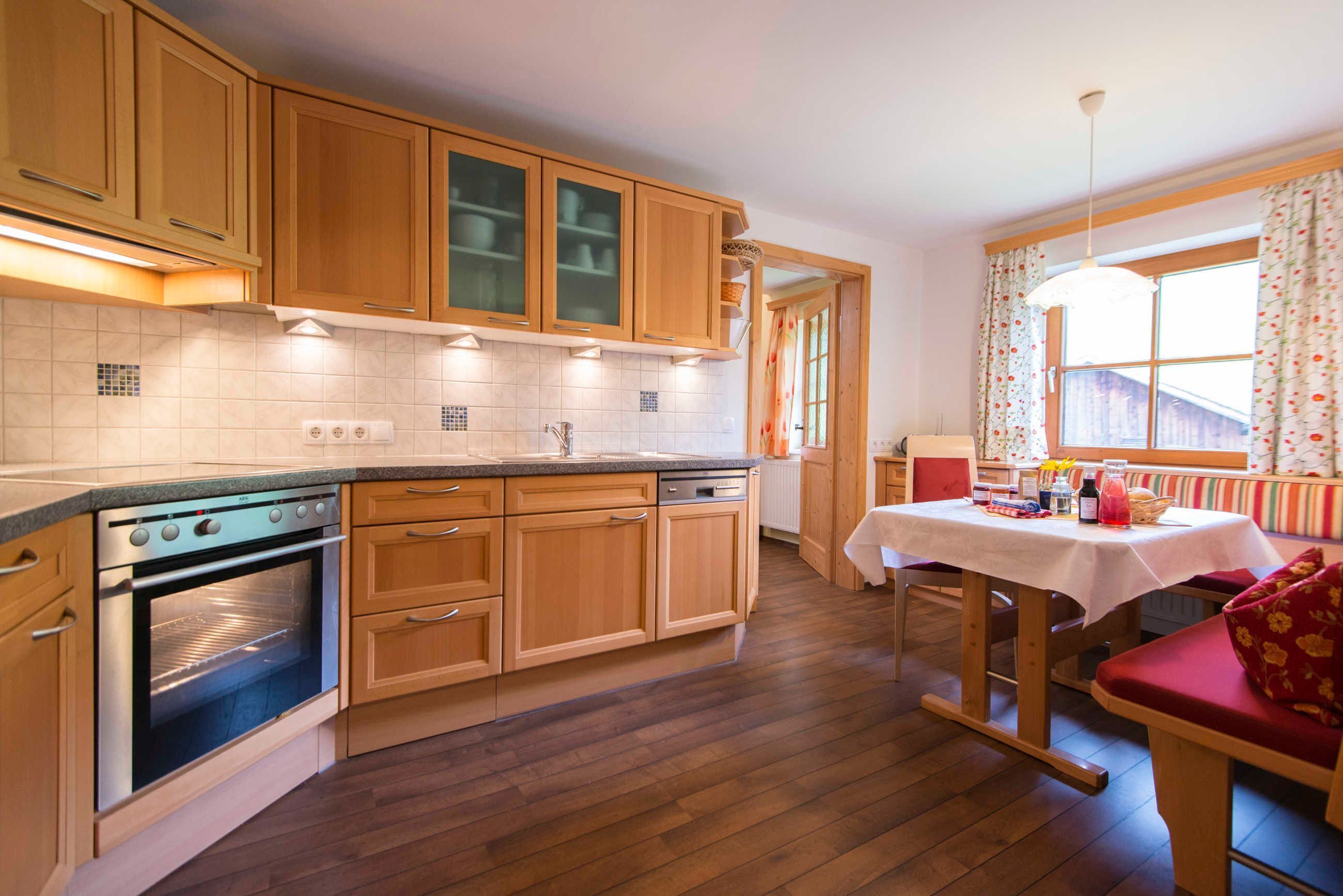 Une cuisine avec des meubles de rangement en bois, un four encastré, un coin repas avec une table et des chaises, et une fenêtre avec des rideaux.