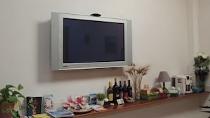 Een flatscreen televisie bevestigd aan een muur boven een houten toonbank met verschillende items zoals wijnflessen, een lamp en decoratieve planten.