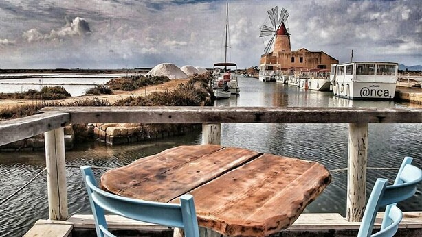 Een houten tafel en twee blauwe stoelen op een steiger, met uitzicht op een windmolen en boten op de achtergrond.