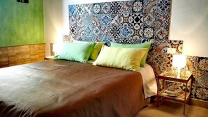 Een keurig opgemaakt bed met een bruine plaid en twee kussens, een nachtkastje met een lamp en een tijdschrift, en een wand met een patroon van tegels.