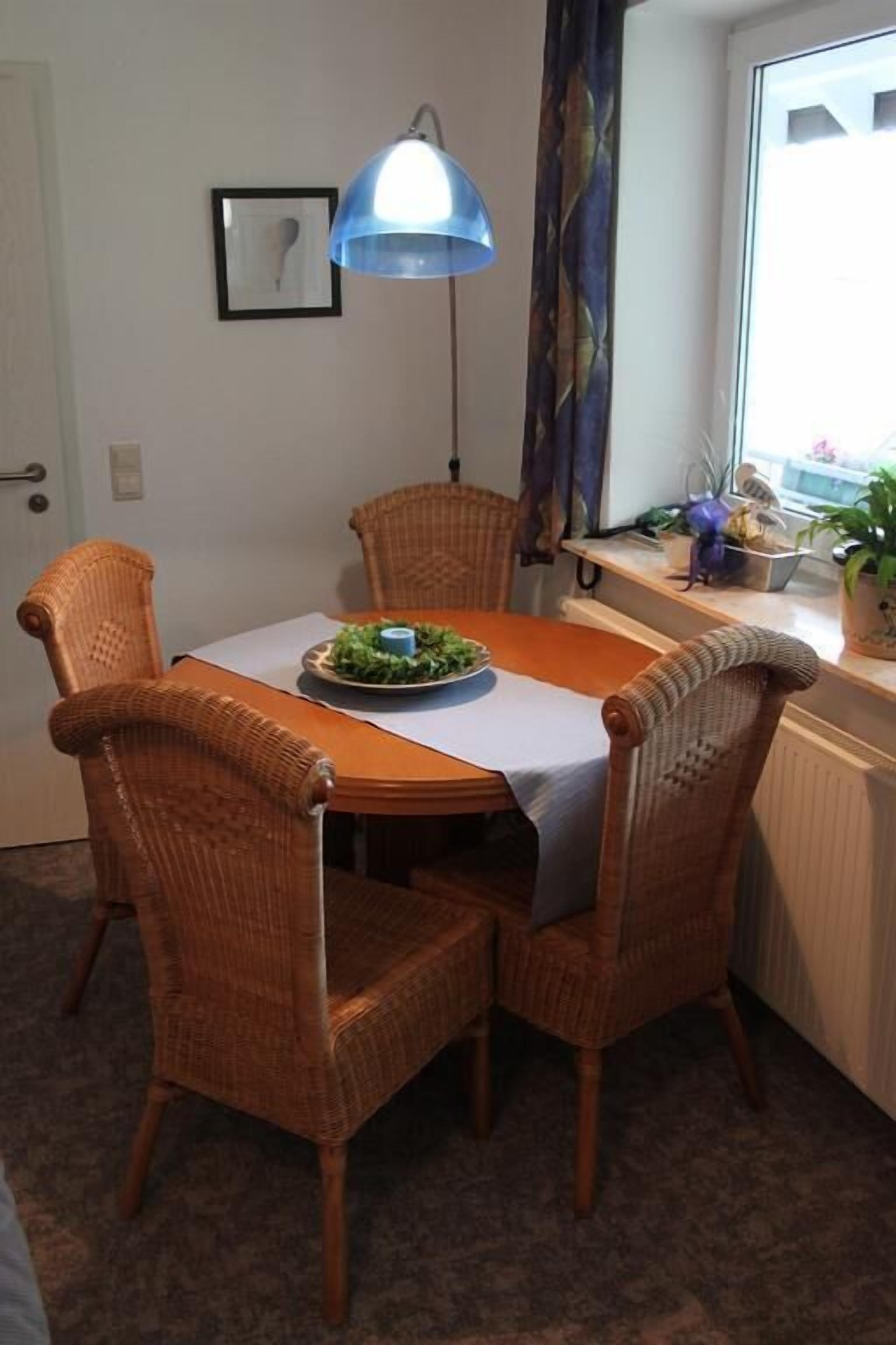 A dining area with wicker chairs, a table with a white tablecloth, a blue lamp, and a window with curtains.