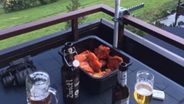 A balcony with a view of a forested landscape, a grill with food, and a table with beer and a bottle. - Ferienwohnung Markus (Schmallenberg)