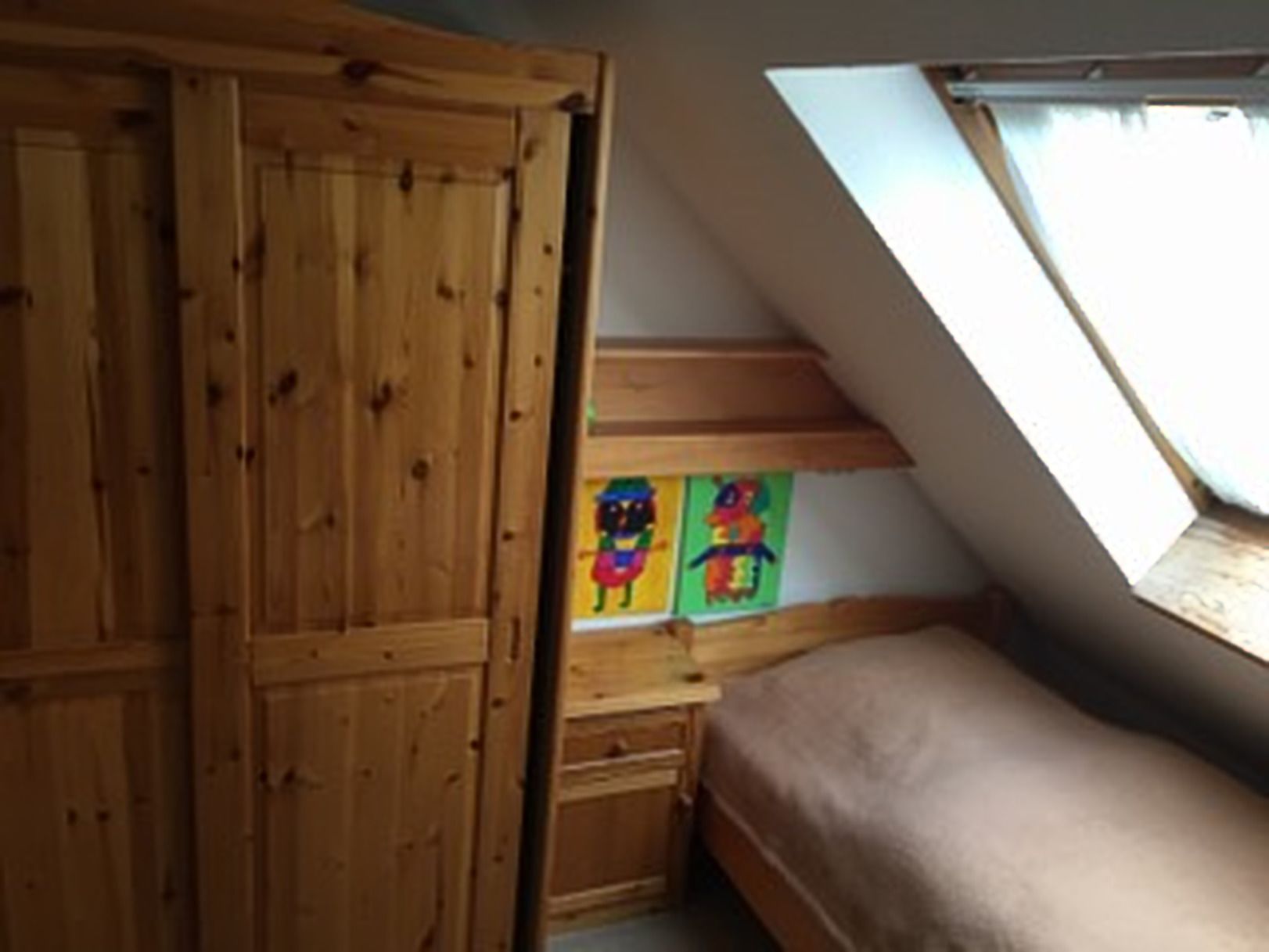 Un dormitorio con un armario de madera, una cama, un escritorio con ilustraciones coloridas y un tragaluz.