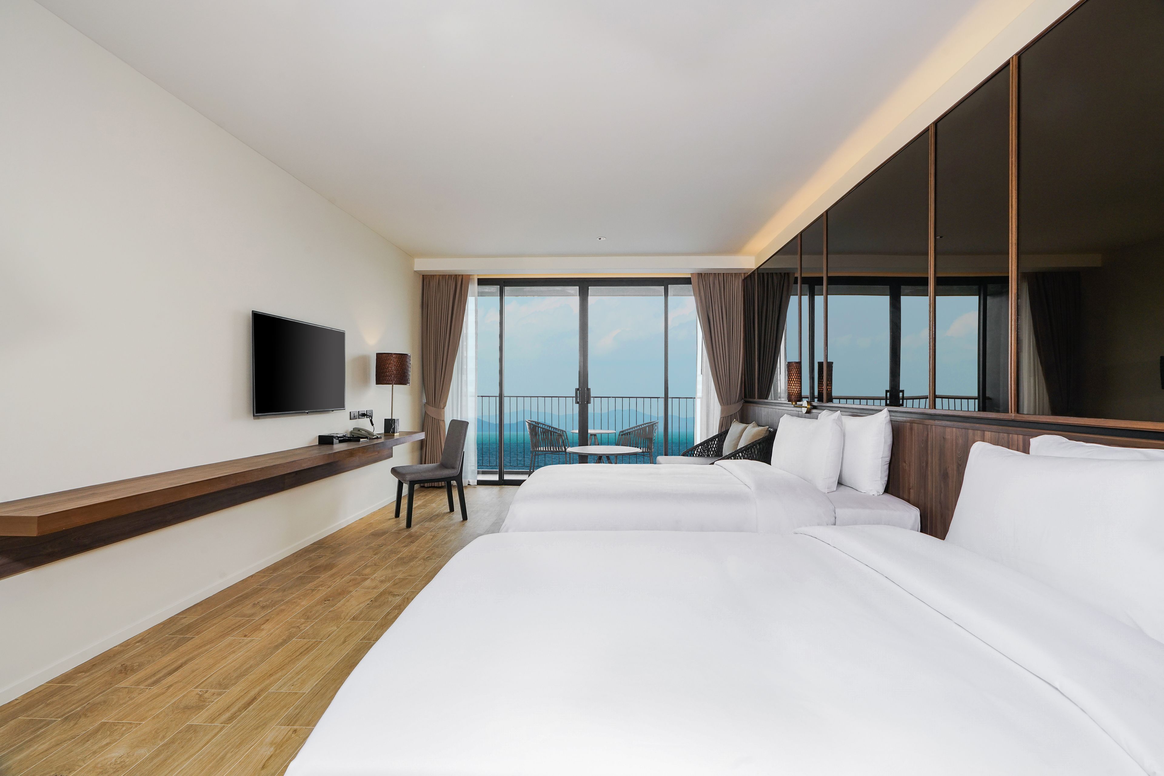A hotel room with a large bed, a TV mounted on the wall, a seating area with a table and chairs, and a balcony with a view.