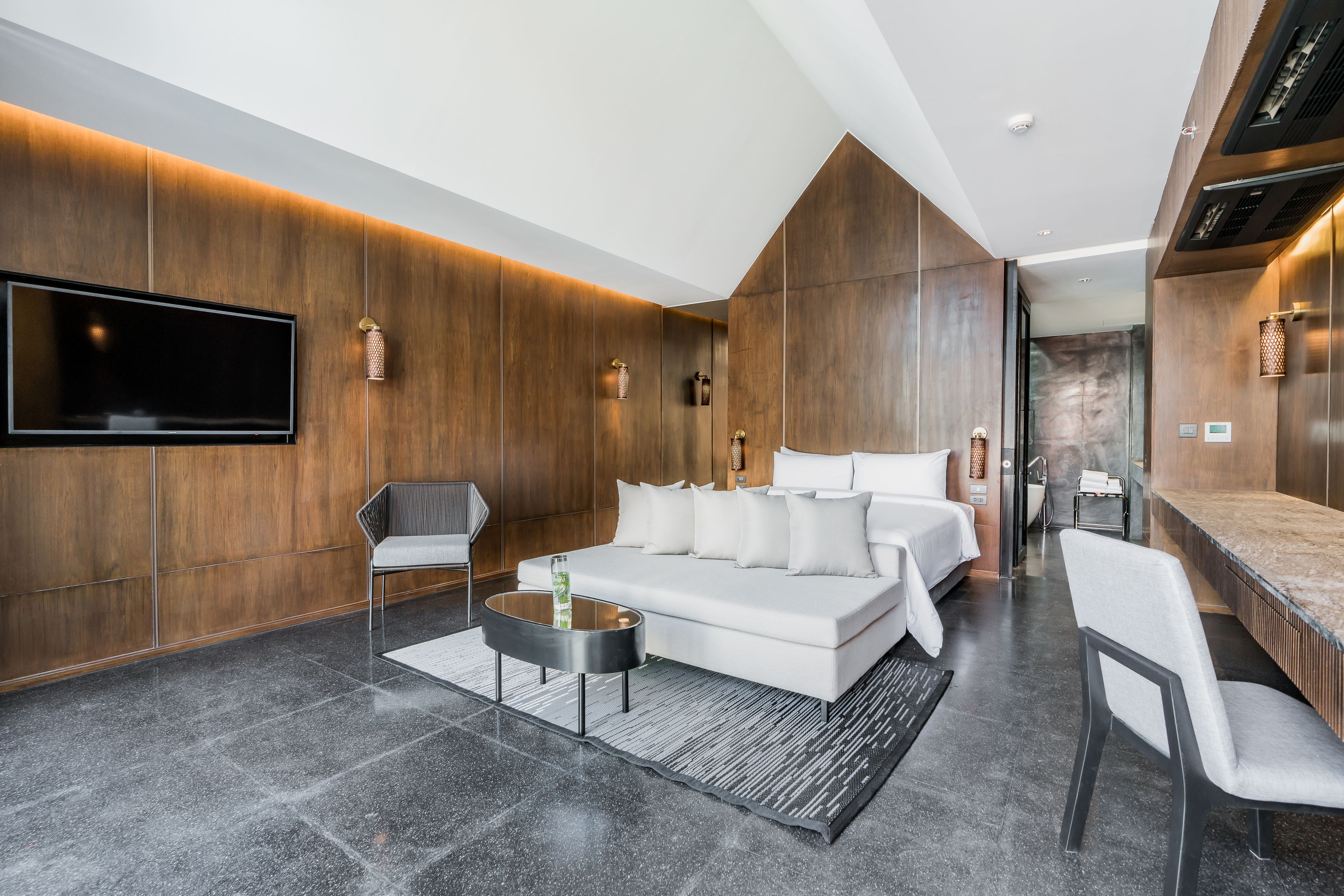 A modern hotel room with a large bed, a sofa, a TV, and a seating area.