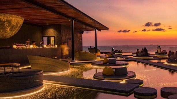 Une piscine à débordement avec des espaces de détente circulaires et un bar, offrant une vue sur la mer au coucher du soleil.