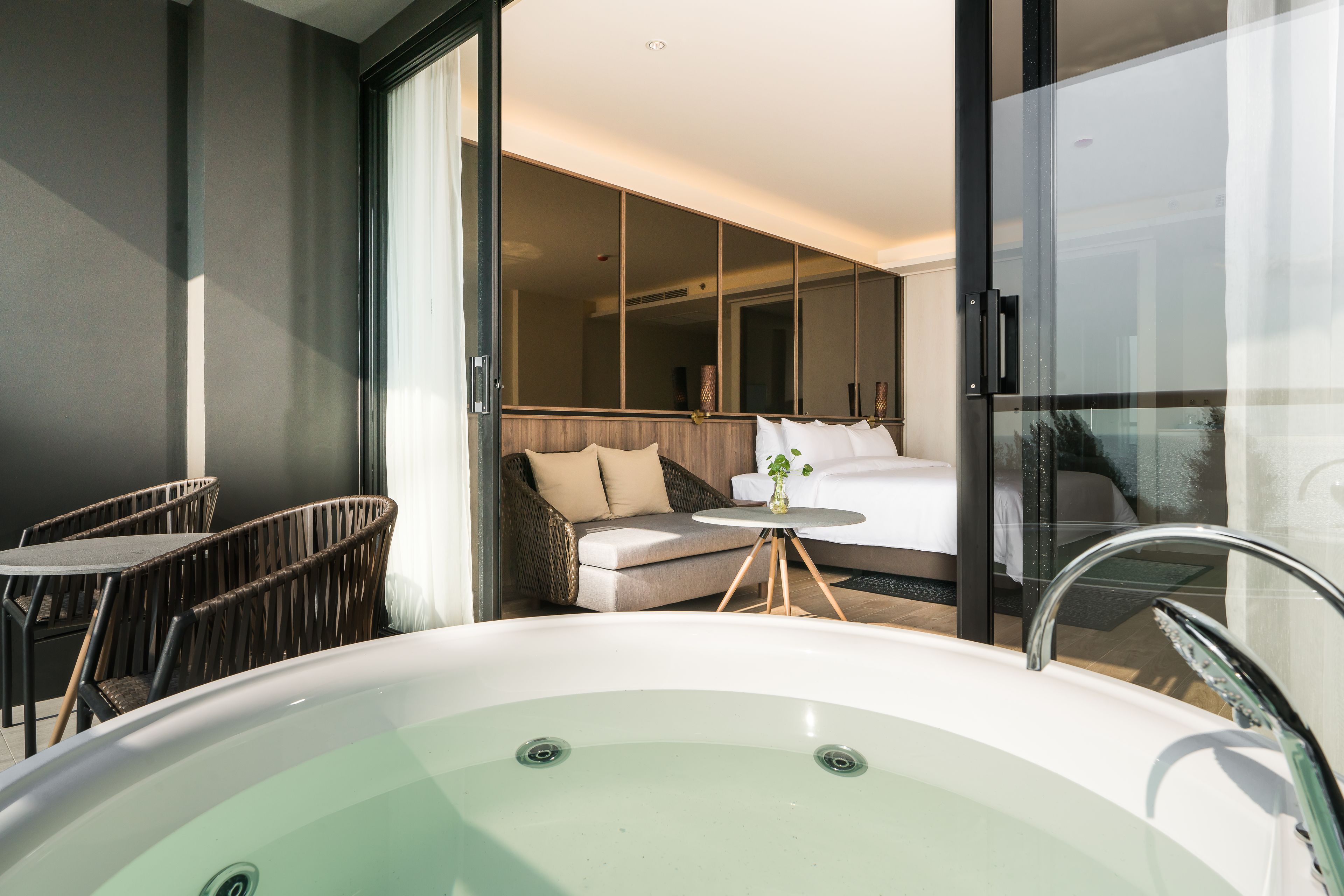 A modern hotel room with a large bathtub, wicker furniture, a sofa, and a glass-enclosed balcony.