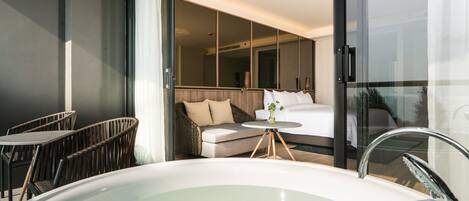A modern hotel room with a large bathtub, wicker furniture, a sofa, and a glass-enclosed balcony.