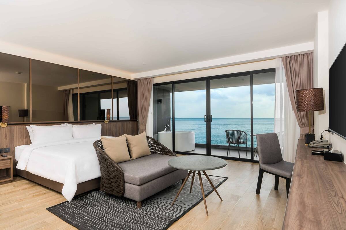 Une chambre d’hôtel moderne avec un grand lit, un canapé, une petite table et une chaise. La chambre dispose d’une grande fenêtre donnant sur la mer.