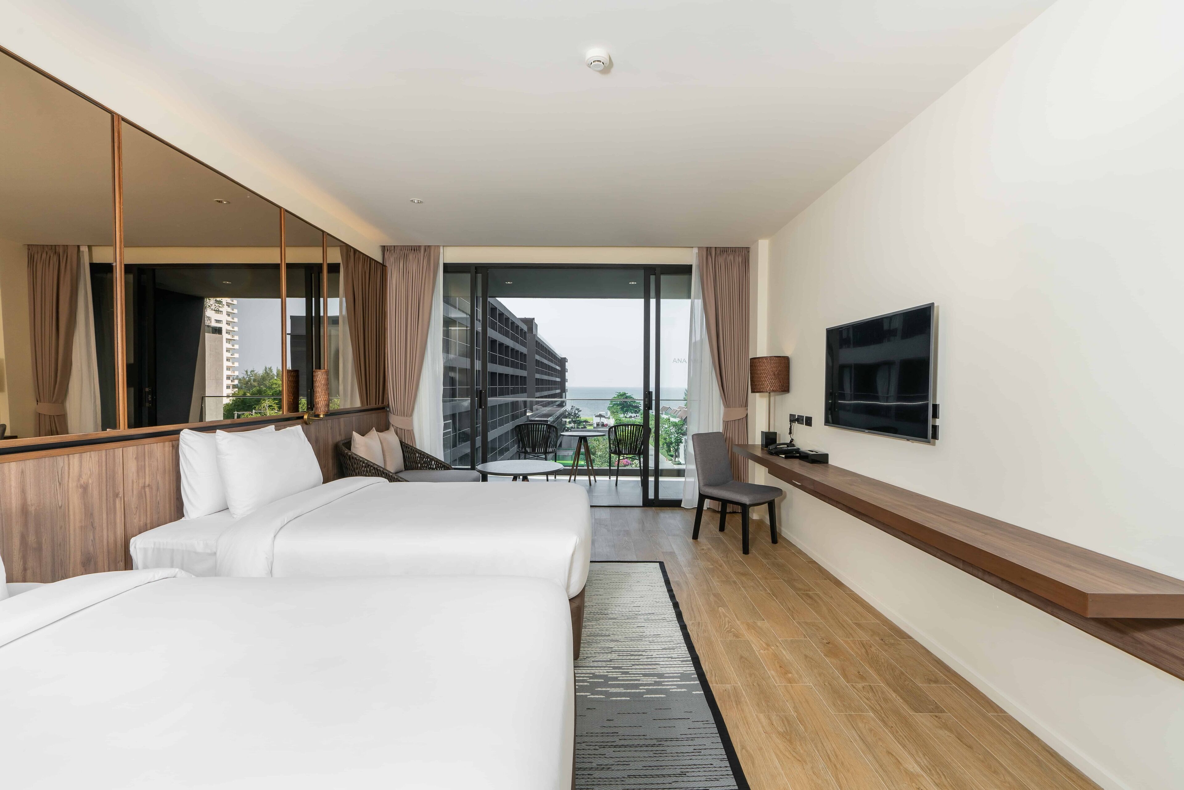Habitación de hotel con dos camas, televisor, balcón con vistas a edificios y zonas verdes, y piso de madera.