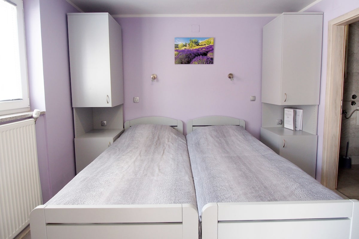 Un dormitorio con dos camas individuales, un armario blanco, una mesita de noche pequeña, un radiador y una ventana.
