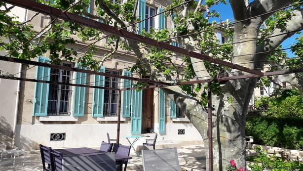 Une terrasse avec du mobilier d’extérieur, un arbre et un bâtiment aux volets bleus.