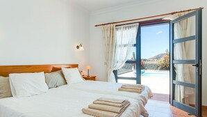 A hotel room with a large bed, a nightstand with a lamp, and a view of a pool and mountains through an open door.