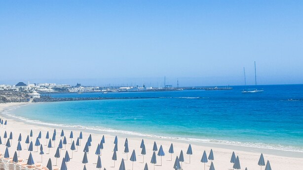 A beach with rows of umbrellas and lounge chairs, a clear blue sea, and a distant cityscape with buildings and wind turbines.
