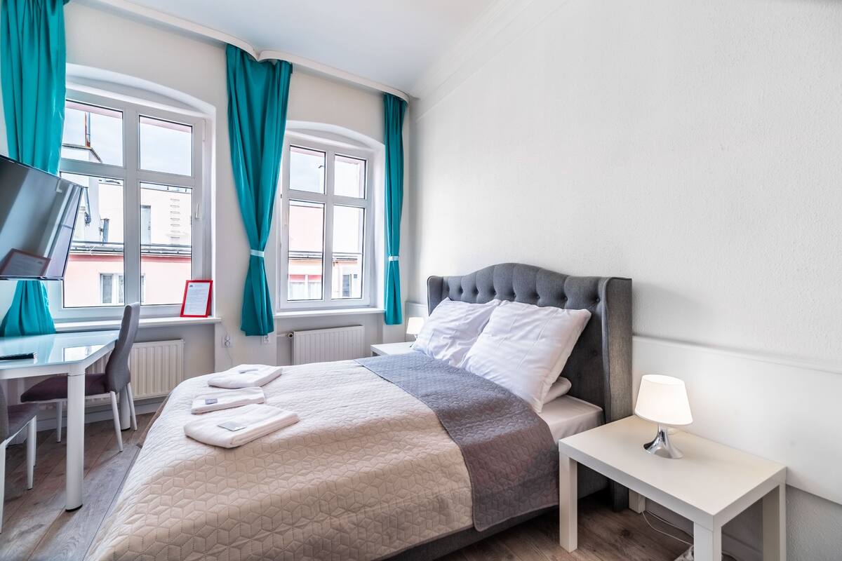 A hotel room with a bed, a desk, a chair, a nightstand, and a window with curtains.
