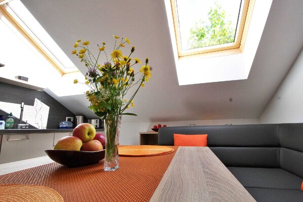 A modern kitchen with a skylight, a dining area with a table and a bowl of fruit, and a gray sofa with an orange cushion.