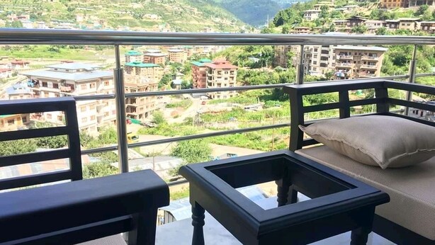 Un balcon doté d’une table et de chaises en verre, offrant une vue sur un village de montagne avec ses bâtiments et sa végétation.