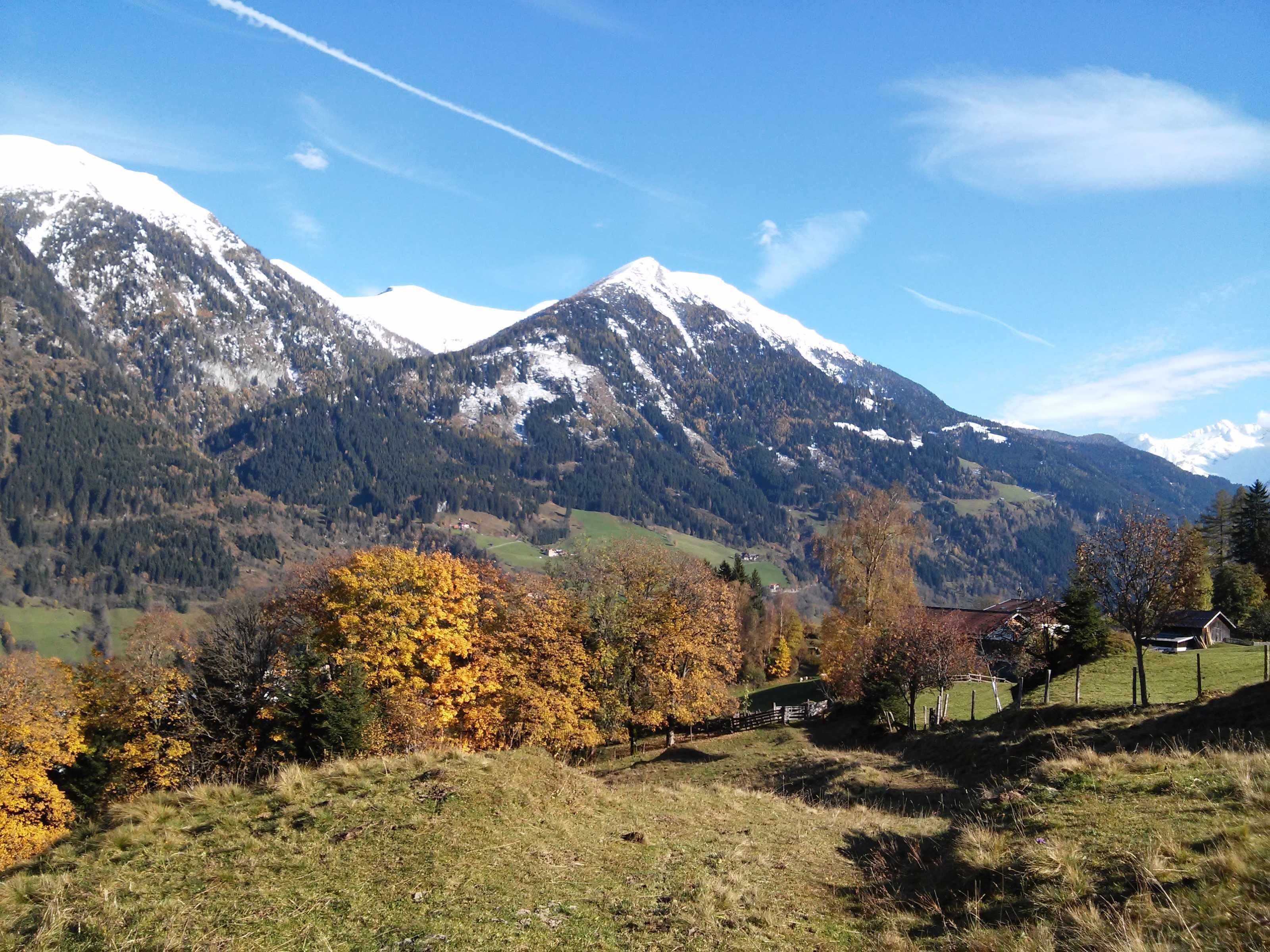 A mountainous landscape with snow-capped peaks, a valley with autumn-colored trees, and a small house in the foreground.