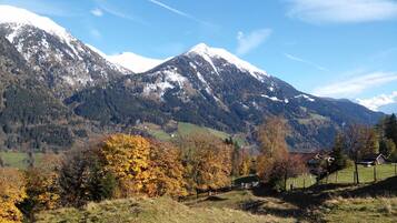 A mountainous landscape with snow-capped peaks, a valley with autumn-colored trees, and a small house in the foreground.