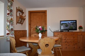 Cozy dining area with a round table and four chairs, featuring a vase with a floral arrangement and a television mounted above a wooden cabinet.