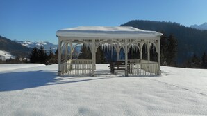 Ein mit Schnee bedeckter Pavillon mit einer hölzernen Bank in der Mitte, umgeben von verschneiter Landschaft und Bergen im Hintergrund.