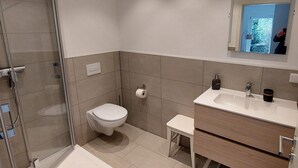 Ein modernes Badezimmer mit Duschkabine aus Glas, Toilette, Waschbecken mit Spiegel und einem kleinen Hocker.