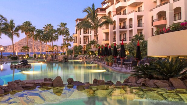 Poolbereich eines Resorts mit Palmen, Liegestühlen und einem Berg im Hintergrund.