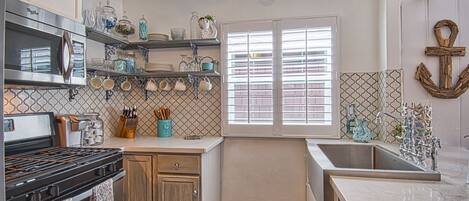 A kitchen with a stove, oven, and microwave. There are shelves with dishes and utensils. A window with blinds is visible.