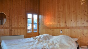 Habitación tipo cabaña de madera con cama, un espejo redondo y ventana con vistas a montañas nevadas.