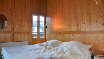 Ein Zimmer in einer Holzhütte mit einem Bett, einem runden Spiegel und einem Fenster mit Blick auf schneebedeckte Berge.
