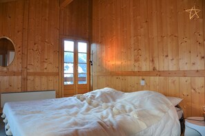 A wooden cabin room with a bed, a round mirror, and a window with a view of snow-covered mountains.