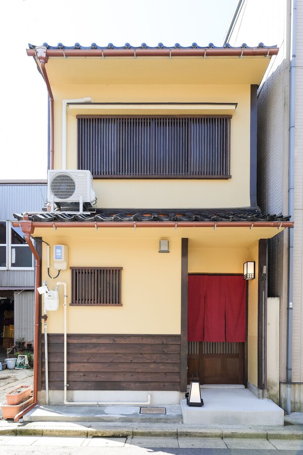 A two-story building with a brown door, a red curtain, and an air conditioning unit on the roof. - Kanazawa no yado Ikedaya (Kanazawa)