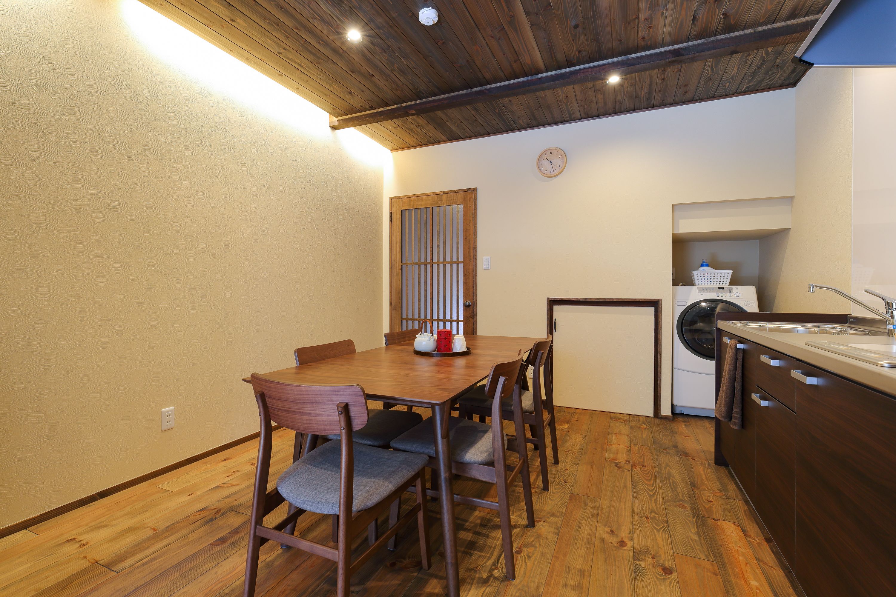 A compact kitchen with wooden flooring, a dining table with chairs, a washing machine, and a clock on the wall.