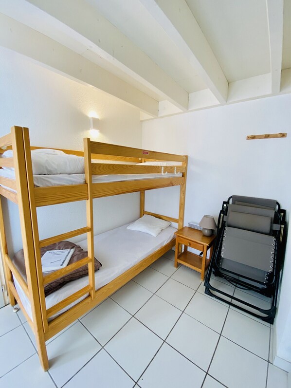 A bunk bed with a small table and a folding chair in a room with white walls and tiled flooring. - Agréable pavillon dans résidence avec piscine (014) (Biscarrosse)
