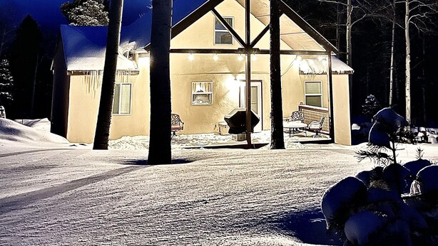 Una casa cubierta de nieve, con un porche delantero y una pequeña terraza, rodeada de altos árboles.
