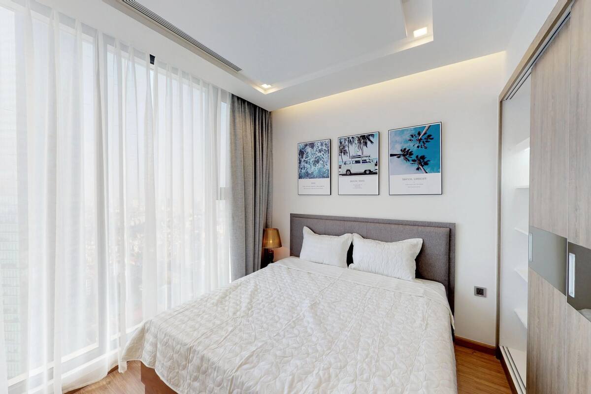 A modern hotel room with a large bed, a nightstand, and a wall-mounted TV. There are three framed pictures on the wall, and the room has large windows with sheer curtains.